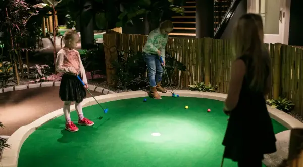 Barn spelar minigolf inomhus i Stockholm. En tropisk miljö med bambustaket och palmer.
