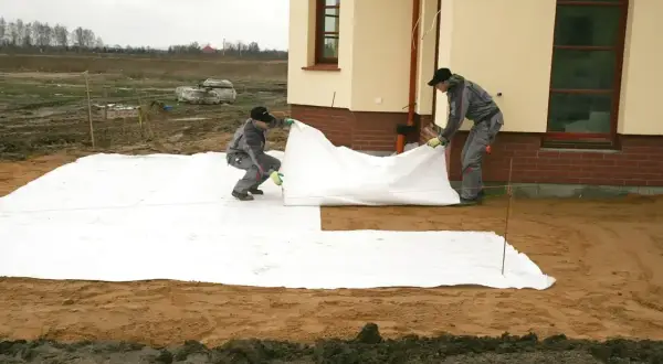 Dwóch robotników rozkłada białą włókninę na placu budowy obok domu.