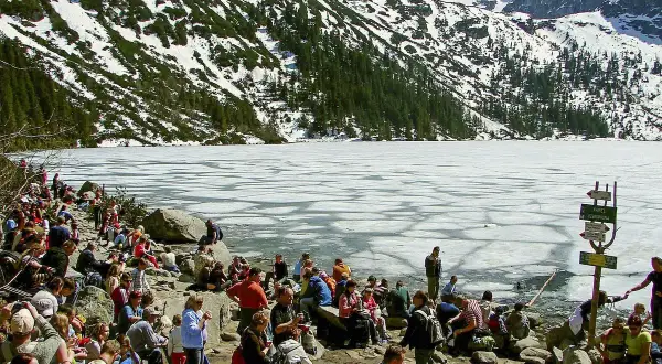 Tłum ludzi odpoczywa nad zamarzniętym jeziorem Morskie Oko, w tle góry pokryte śniegiem i lasem. Palenica Białczańska w oddali.