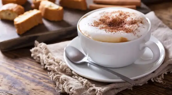 Un cappuccino senza latte, con schiuma soffice e cacao, accompagnato da biscotti secchi e cannella su un tavolo di legno rustico.