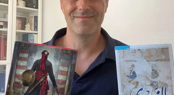 Juan de Granada, sonriente, sostiene dos libros. Uno muestra un guerrero con armadura y el otro, una escena histórica.