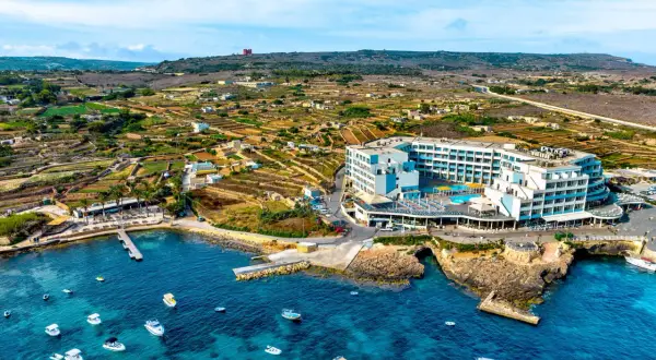 Malta last minute tanio: hotel z basenem nad turkusowym morzem, otoczony tarasami i łodziami. W tle malownicze, tarasowe pola i wzgórza.