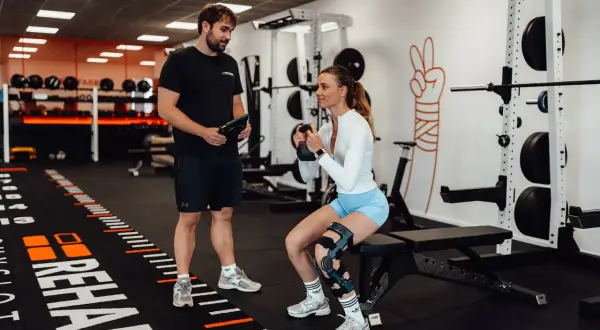 Mann mit Tablet beobachtet Frau beim Kniebeugen-Training mit Kettlebell. Meniskus Training statt OP ist hier das Ziel.