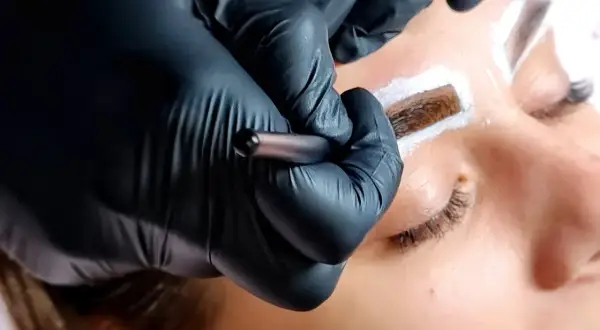 Stylist w czarnych rękawiczkach aplikuje hennę pudrową na brwi klientki.