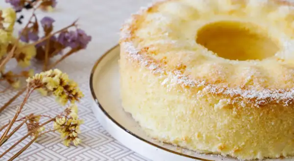 Delizioso chiffon cake ricetta originale americana, leggero e soffice, spolverato di zucchero a velo, con fiori secchi sullo sfondo.