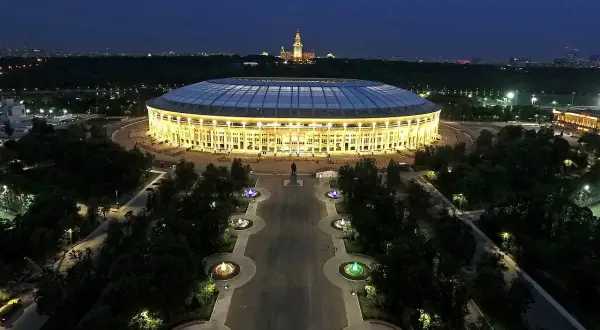 Oświetlony moskiewski stadion nocą, z aleją fontann i pomnikiem w tle.