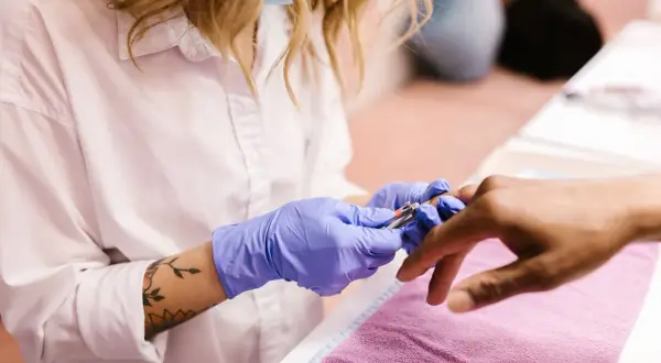 Manicurzystka w niebieskich rękawiczkach precyzyjnie usuwa skórki wokół paznokci klienta za pomocą cążków.