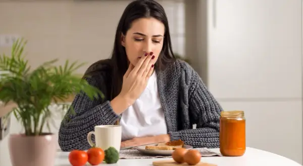 Donna con nausea e debolezza alle gambe si copre la bocca, seduta a tavola con colazione.