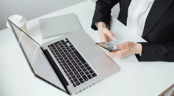 Kobieta w czarnej marynarce trzyma telefon, obok laptop i kubek. Zastanawia się, jak zaadresować pismo do adwokata.