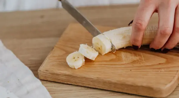 Eine Hand schneidet eine Banane in Scheiben auf einem Holzbrett. Perfekte Energie für das Training.
