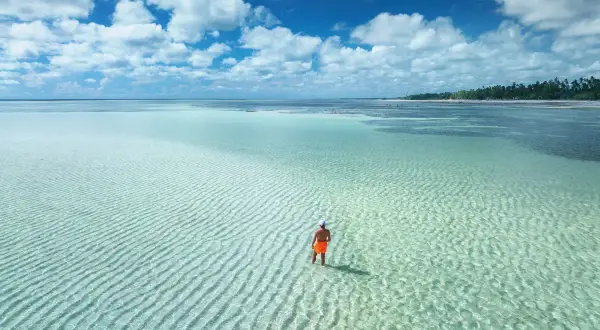 Człowiek w pomarańczowych spodenkach stoi w płytkiej, turkusowej wodzie. Zastanawia się, ile się leci na Zanzibar, by dotrzeć do tego raju.
