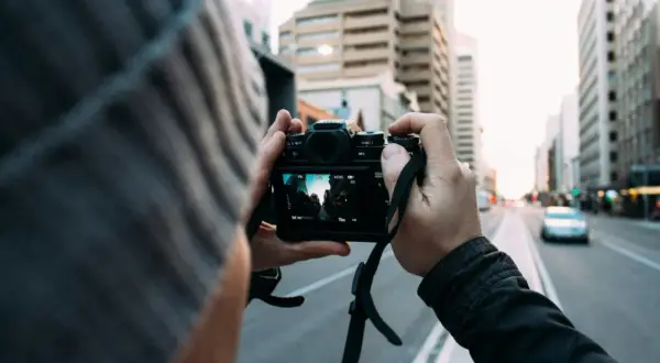 Fotografia reportażowa uchwycona przez osobę w czapce, trzymającą aparat i fotografującą miejski krajobraz z budynkami i ulicą.