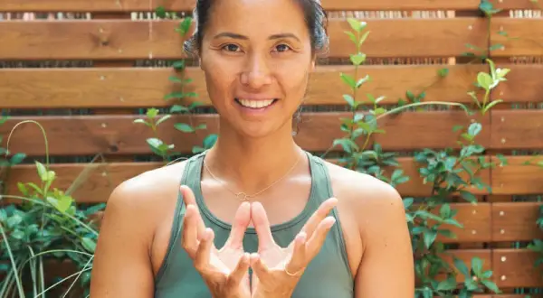 Mujer sonriente con las manos en posición de mudra, rodeada de vegetación y una valla de madera.