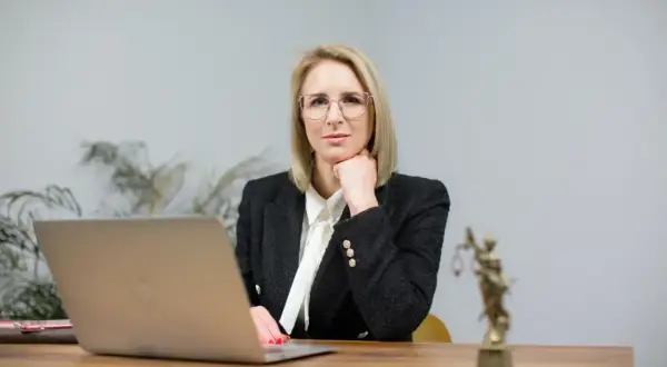 Kobieta w okularach i żakiecie siedzi przy biurku z laptopem i figurką Temidy.