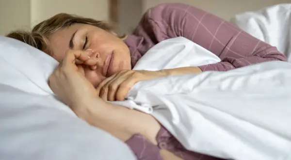 En kvinna sover 6 timmar i en säng med vita lakan och en lila rutig pyjamas. Hon håller handen mot ansiktet.
