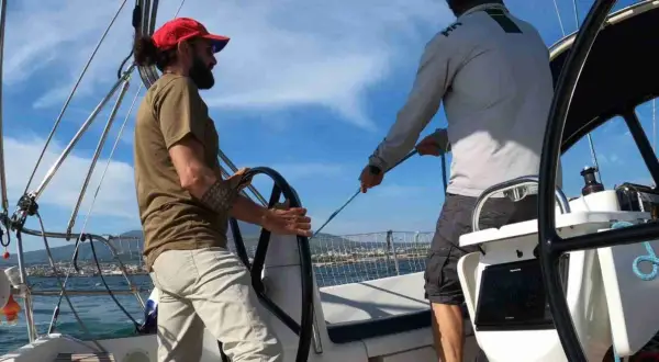 Due uomini su una barca a vela sport, uno al timone, l'altro che regola le cime. Cielo azzurro e mare calmo.