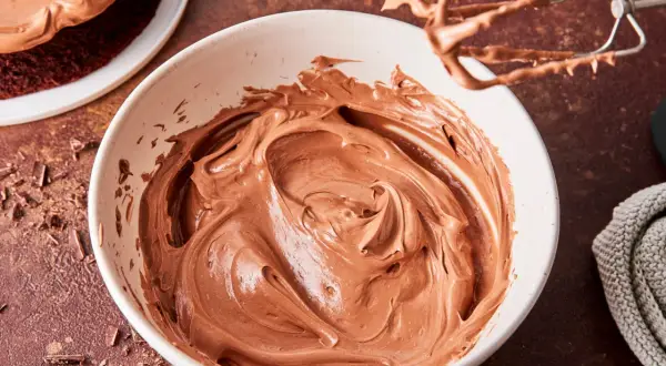 Schoko-Buttercreme in einer Schüssel, bereit zum Verzieren eines Kuchens. Ein Handmixer mit Schoko-Buttercreme hängt darüber.