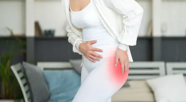 A woman in white clothing holds her hip, which is highlighted in red, indicating pain. She might be wondering, "why do my hips hurt when I sleep?