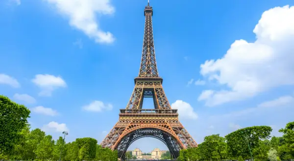 The Eiffel Tower stands tall against a bright blue sky with fluffy white clouds, surrounded by lush green trees and a manicured lawn.