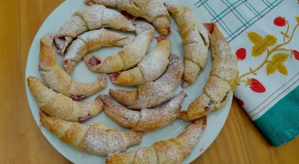 Pyszne, domowe rogaliki z marmoladą, posypane cukrem pudrem, ułożone na talerzu obok ręcznika z truskawkami.