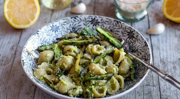 Un plat de pâtes aux asperges, garni de parmesan râpé, avec des citrons et de l'ail en arrière-plan.
