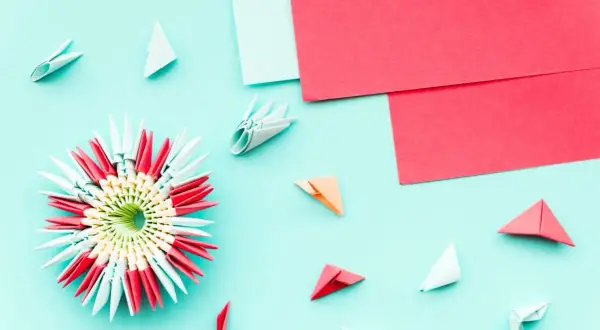 Pyssel med papper: en färgglad 3D-origamiblomma omgiven av små vikta pappersdelar och ark i rött och ljusblått.