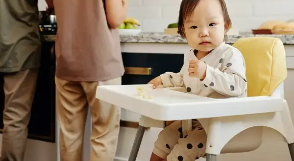 Małe dziecko siedzi w krzesełku dla dziecka, jedząc kawałki banana. W tle widać dwie osoby przygotowujące posiłek w kuchni.