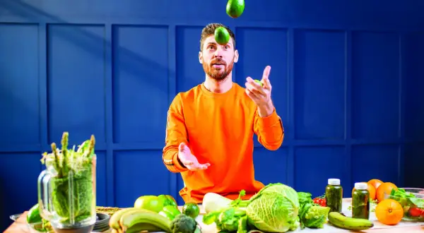 Un homme jongle avec des avocats, entouré de légumes frais. Une table garnie de fruits et légumes suggère une alimentation saine pour gérer l'acide urique.