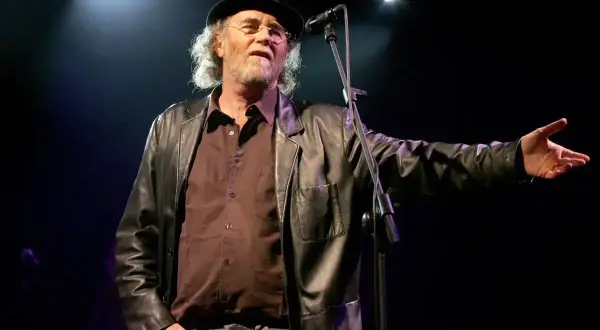 Francesco De Gregori, con cappello e giacca di pelle, canta sul palco. I suoi testi, come "Adelante Adelante", risuonano nell'aria.
