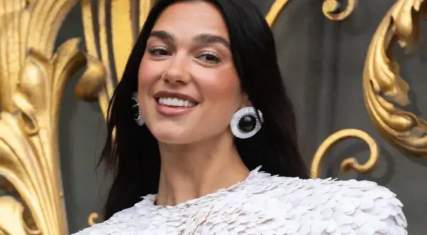 Uśmiechnięta Dua Lipa w białej kreacji, z dużymi kolczykami. Jej albańskie pochodzenie dodaje jej uroku.