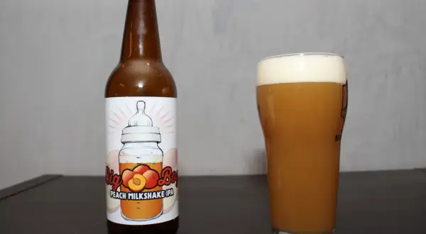 Butelka i szklanka z brzoskwiniowym milkshake IPA. Etykieta przedstawia butelkę z smoczkiem i brzoskwiniami.