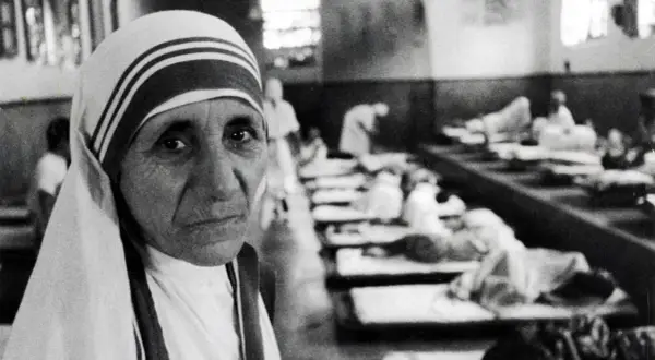 Mutter Teresa, deren Herkunft in Skopje liegt, blickt nachdenklich in die Kamera. Im Hintergrund liegen Kranke in Betten.