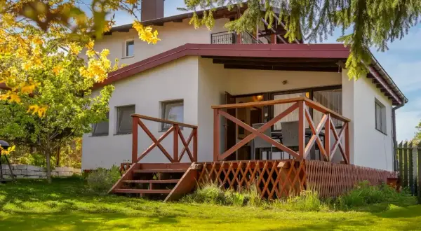 Bungalow z drewnianym tarasem i schodkami, otoczony zielonym ogrodem i kwitnącym drzewem.