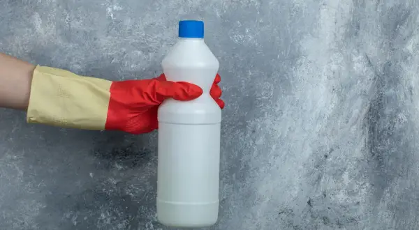 Mano con guanto rosso e giallo tiene una bottiglia bianca di candeggina, pronta per pulire il cemento.