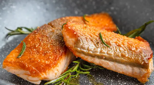 Zwei saftige Lachsfilets mit knuspriger Haut, perfekt gebraten, liegen in einer Pfanne mit Rosmarin und Öl.
