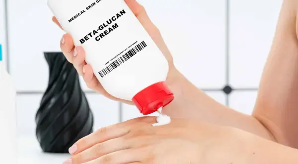Eine Hand hält eine Tube "Beta-Glucan Cream" und gibt eine kleine Menge auf die andere Hand. Die Creme verspricht eine positive **Beta-Glucan Wirkung Haut**.