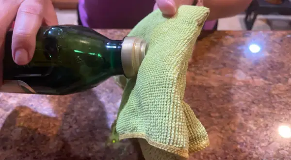 A hand pours olive oil from a green bottle onto a green microfiber cloth, suggesting it's the best home remedy to clean stainless steel.
