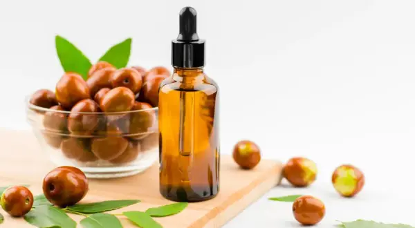 Olio di jojoba a cosa serve? Un flacone ambrato con contagocce accanto a una ciotola di frutti di jojoba e foglie verdi.