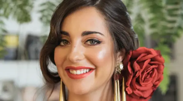 Peinados para feria pelo corto: mujer sonriente con rosa roja en el cabello, pendientes dorados y maquillaje festivo.
