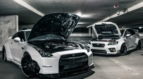 Deux voitures de sport blanches, capots ouverts, révélant leurs moteurs. Ces chevaux fiscaux, prêts à rugir, sont garés dans un parking sombre.