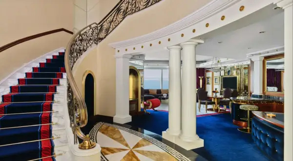 Luksusowe wnętrze z marmurowymi schodami, złotymi zdobieniami i niebieskim dywanem. To widok, który może towarzyszyć pobytowi w Burj Al Arab, gdzie burj al arab cena za noc odzwierciedla tę elegancję.
