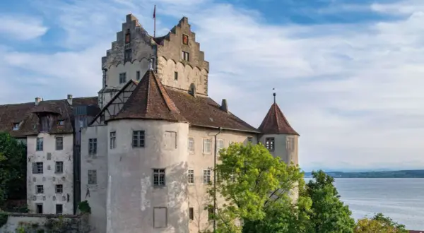 Das Meersburg Castle thront über dem Bodensee, umgeben von grünen Bäumen und unter einem blauen Himmel mit Wolken.