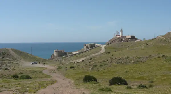 Un camino de tierra lleva al restaurante El Faro Cabo de Gata, con el mar azul y un faro en la colina.