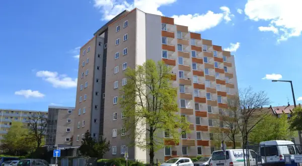 Wysoki blok mieszkalny, który stanowi spadek po rodzicach, z balkonami i zielonym drzewem przed nim.