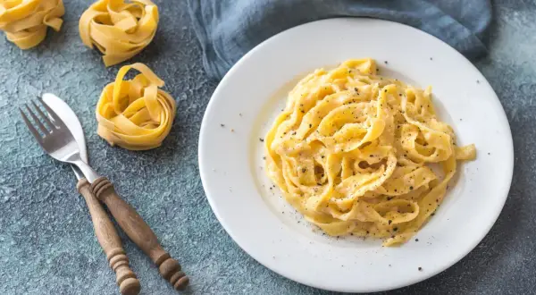 Un plat de fettuccine alfredo crémeux, saupoudré de poivre noir, avec des pâtes crues et une fourchette en bois à côté.