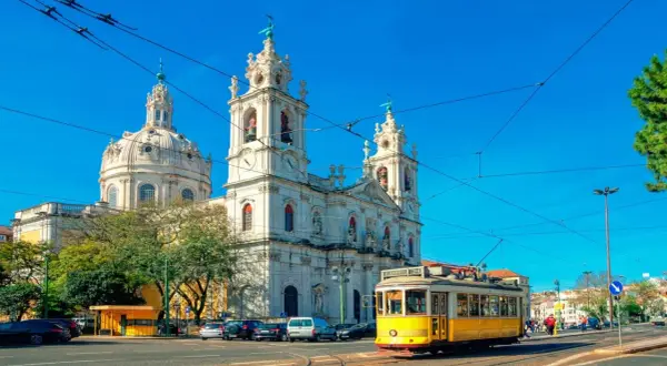 Un tranvía amarillo cruza frente a una iglesia blanca con cúpulas y torres, reflejando cómo se vive en Portugal con su encanto histórico y transporte icónico.