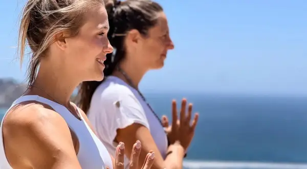 Dos mujeres practican yoga frente al mar, con las manos juntas en gesto de namasté.