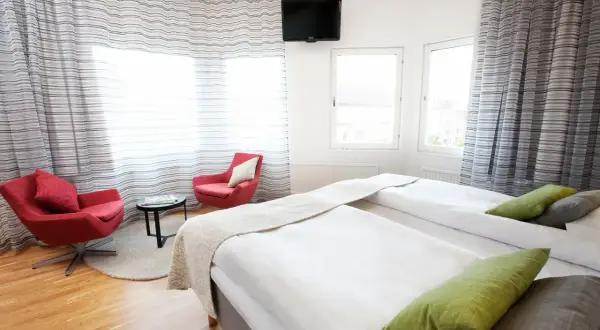Ett modernt hotellrum med två sängar, röda fåtöljer och utsikt över en plaza i Borås.