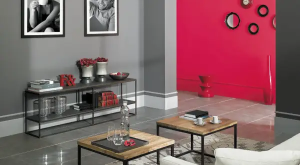 Nowoczesny salon z szarymi ścianami, na których wiszą czarno-białe zdjęcia. Obok, jaskrawoczerwona ściana z dekoracjami. To dowód, jakie kolory pasują do szarego.