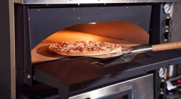Une pizza garnie est en cours de cuisson dans un four professionnel, sur une pelle à pizza. La chaleur intense du four promet une cuisson parfaite.